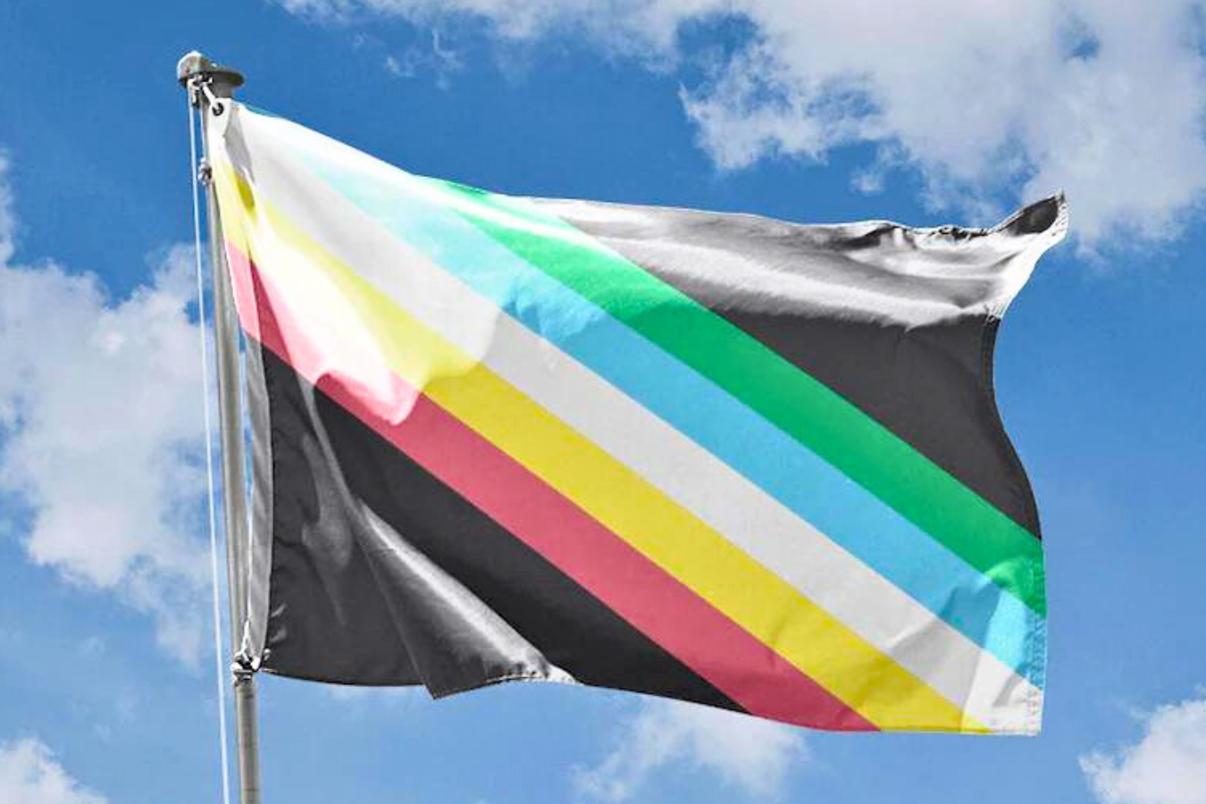 Frente a un cielo azul nublado aparece una bandera con franjas diagonales, rojas, amarillas, blancas, azules y verdes sobre un fondo negro carbón.