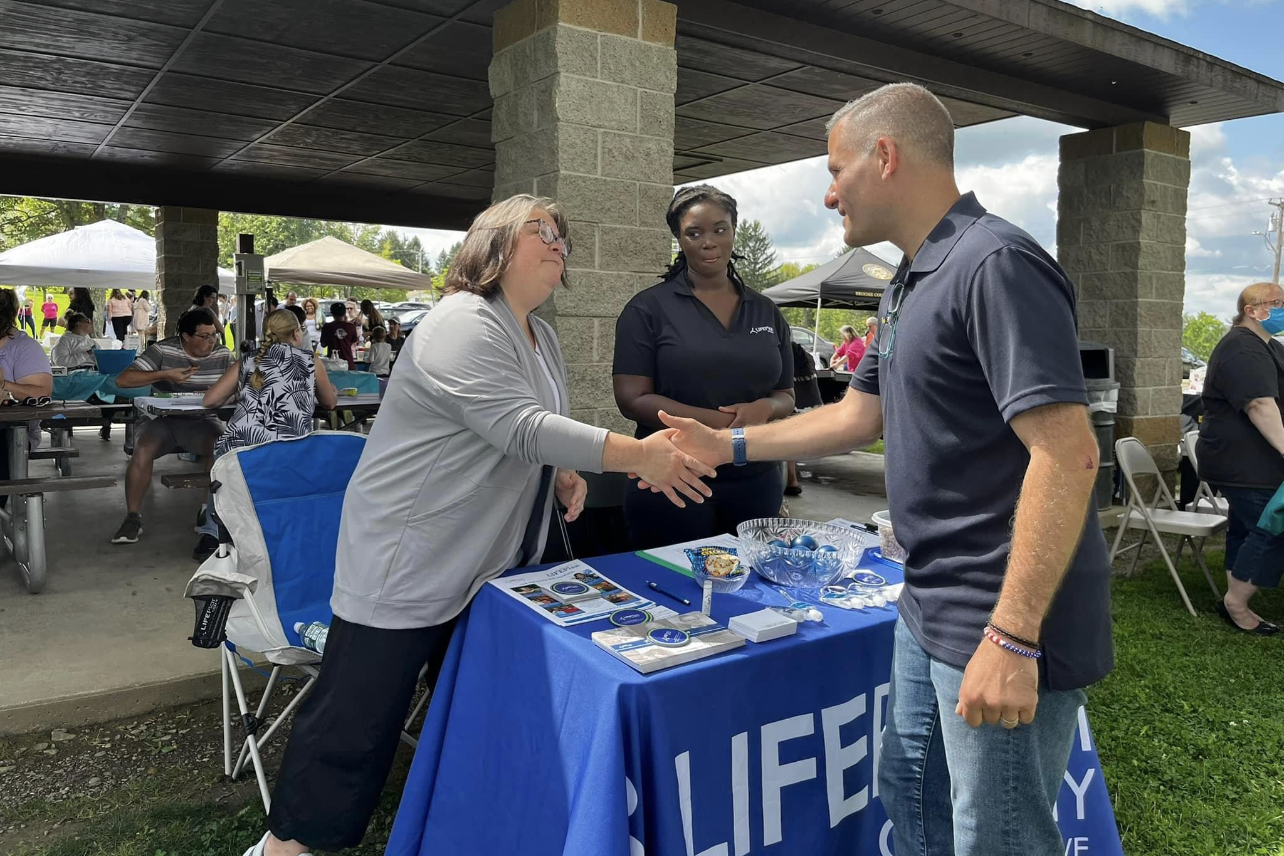A man in a polo shirts shakes hands with two women in from of a LIFEPlan promotional table.