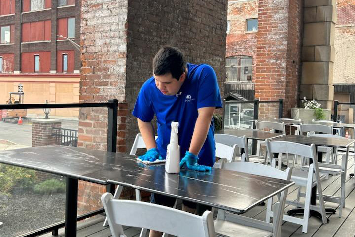 A young man in a blue polo cleans a table on a deck.