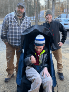 Two men stand on a trail behind a wheelchair-like device with a younger man seated in it.