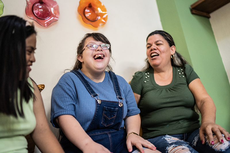 Two women sit on either side of a young woman with a disability who is laughing.