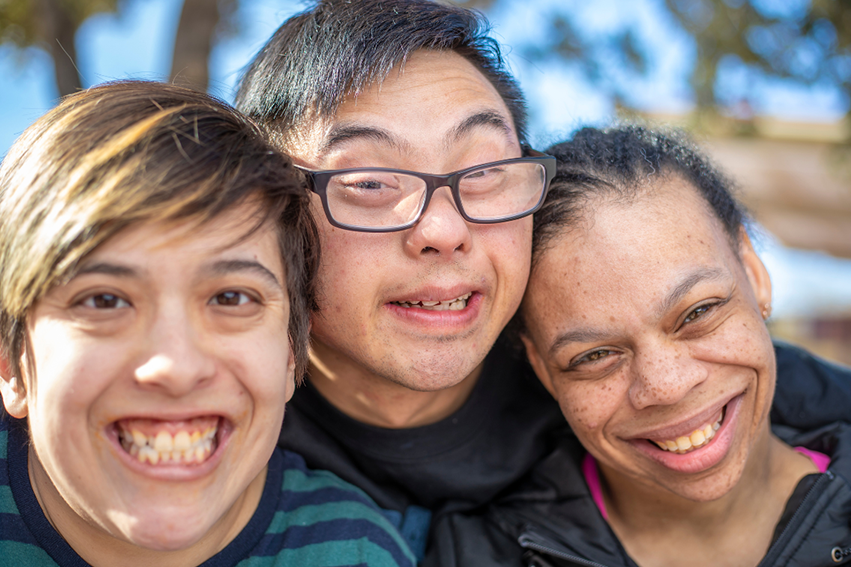 Three young people with disabilities are huddled together with smiles.